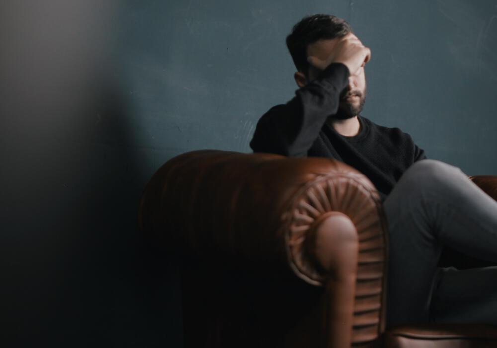 Man sitting on brown couch with his hand covering his face, looking distressed